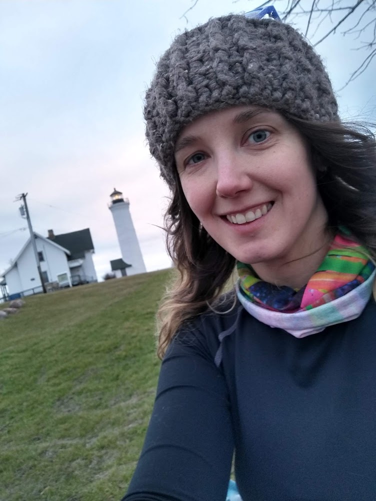 A brown haired white woman stands downhill from a white lighthouse and outbuilding